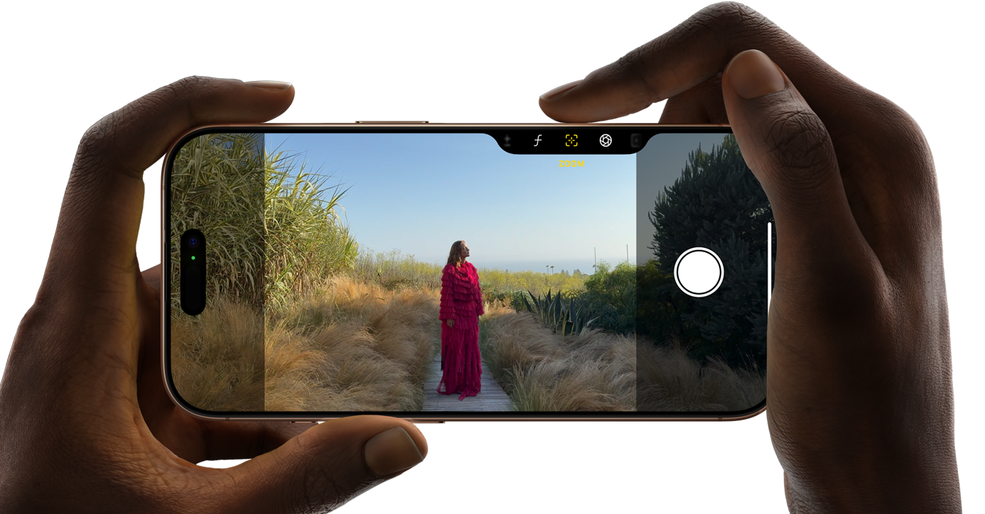 To hænder der holder en iPhone 16 pro vandret i hver deres ende, mobilens skærm er inde på kameraret, hvor man kan se en kvinde i et lyserødt outfit, stående på en træ sti omringet af natur, kvindes øjne er lukket med hendes hage lidt skråt op til højre